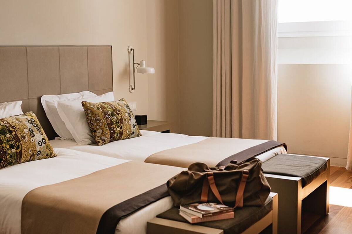 A hotel room with two beds, a bedside table, a bench, and a window with curtains.