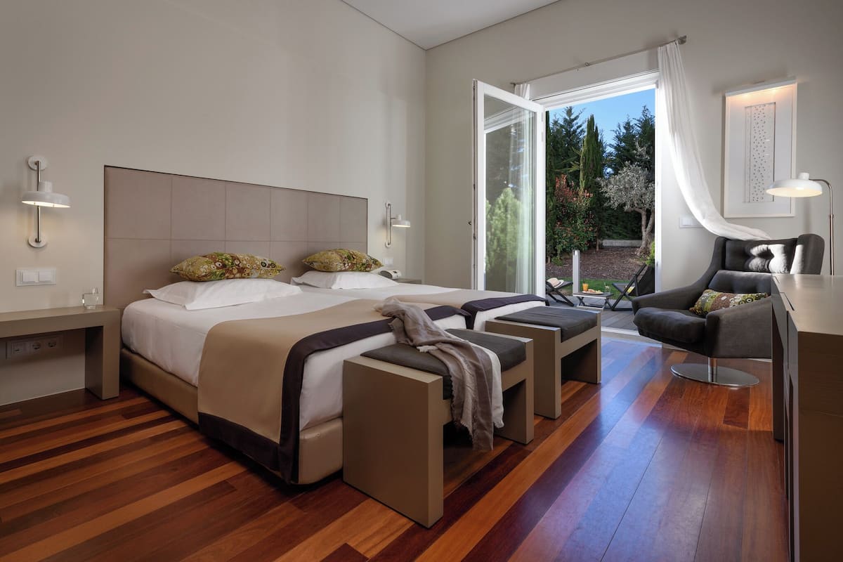 A modern hotel room with a large bed, a desk, and a chair. There is a sliding glass door leading to a balcony with a view of trees and a garden.