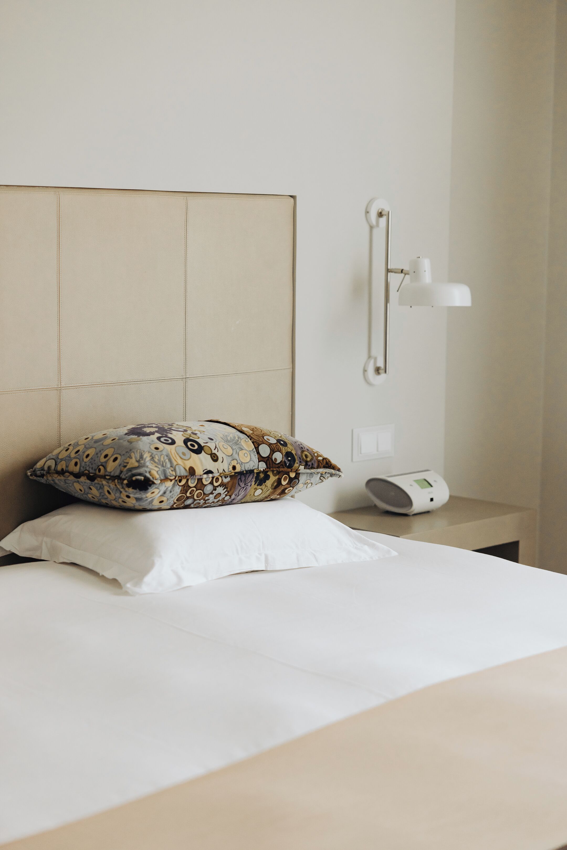 Una cama bien tendida con almohadas estampadas y lisas, una mesita de noche con lámpara y radio, y una lámpara de pared.