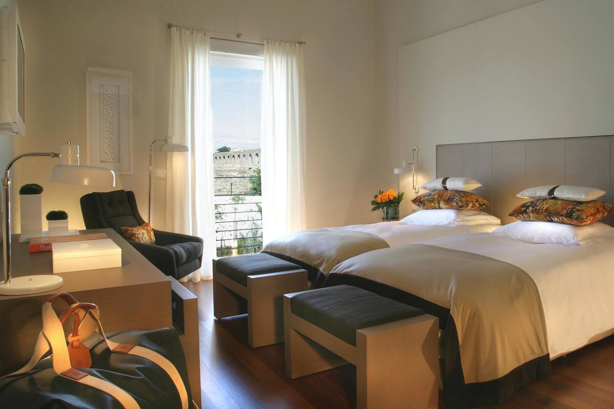 A hotel room with two beds, a desk with a lamp, a chair, a small table, and a view of a historic building through the window.