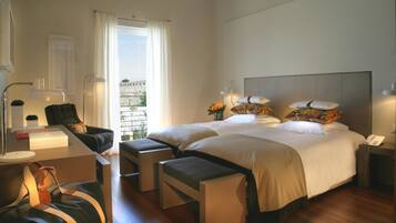 Habitación de hotel con dos camas, un escritorio con lámpara, una silla, una mesita con un florero y vista a un muro de piedra a través de la ventana.