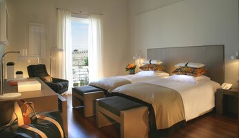 Habitación de hotel con dos camas, un escritorio con lámpara, una silla, una mesita con un florero y vista a un muro de piedra a través de la ventana.