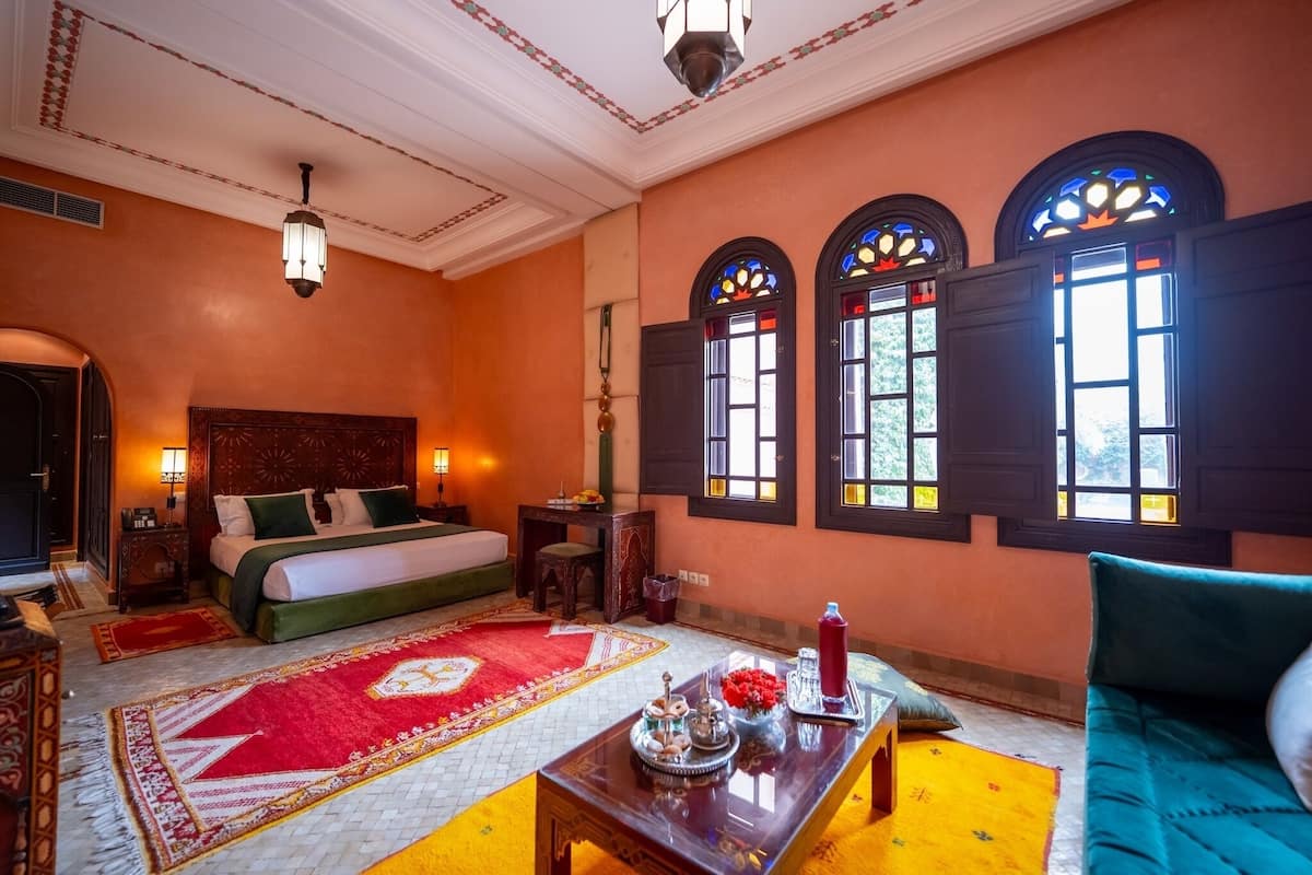 Quarto de hotel com cama, sofá, mesa com objetos decorativos, tapete vermelho e branco, e três janelas com vitrais.