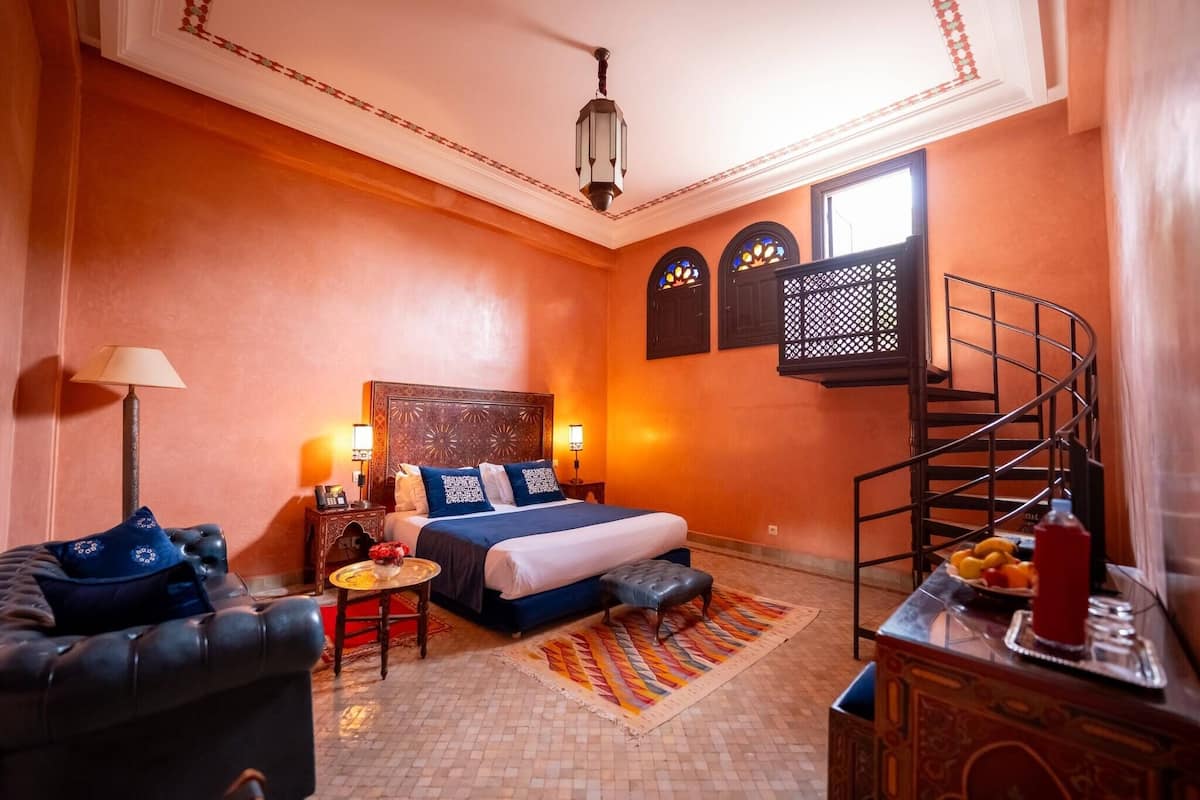 Quarto de hotel com cama, sofá, mesa pequena, luminária, tapete decorativo, escada e prateleira na parede com frutas.