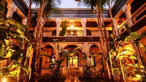 A courtyard with a central fountain, surrounded by palm trees and plants, with a building featuring arches and balconies in the background. - Al Fassia Aguedal (Marrakech)