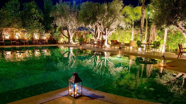 A nighttime pool area with lounge chairs and tables, surrounded by trees and illuminated by ambient lighting. - Al Fassia Aguedal (Marrakech)