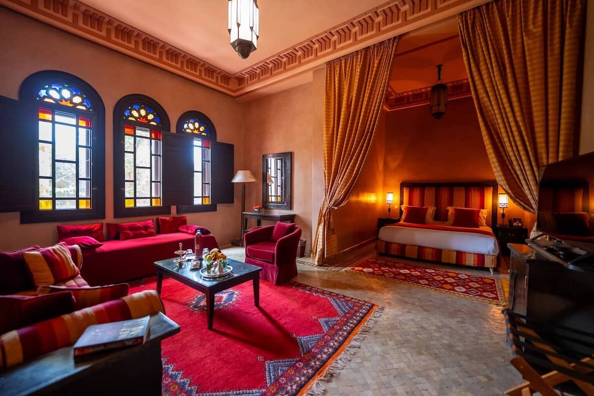 Um quarto acolhedor com um sofá vermelho, uma mesa de centro e uma televisão. Há três janelas com vitrais, uma cortina vermelha e um tapete estampado.