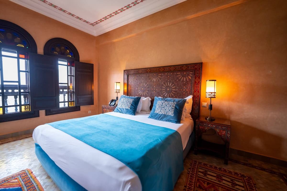 Quarto de hotel com uma cama grande, duas luminárias de cabeceira, um painel de cabeceira estampado e uma janela com persianas.