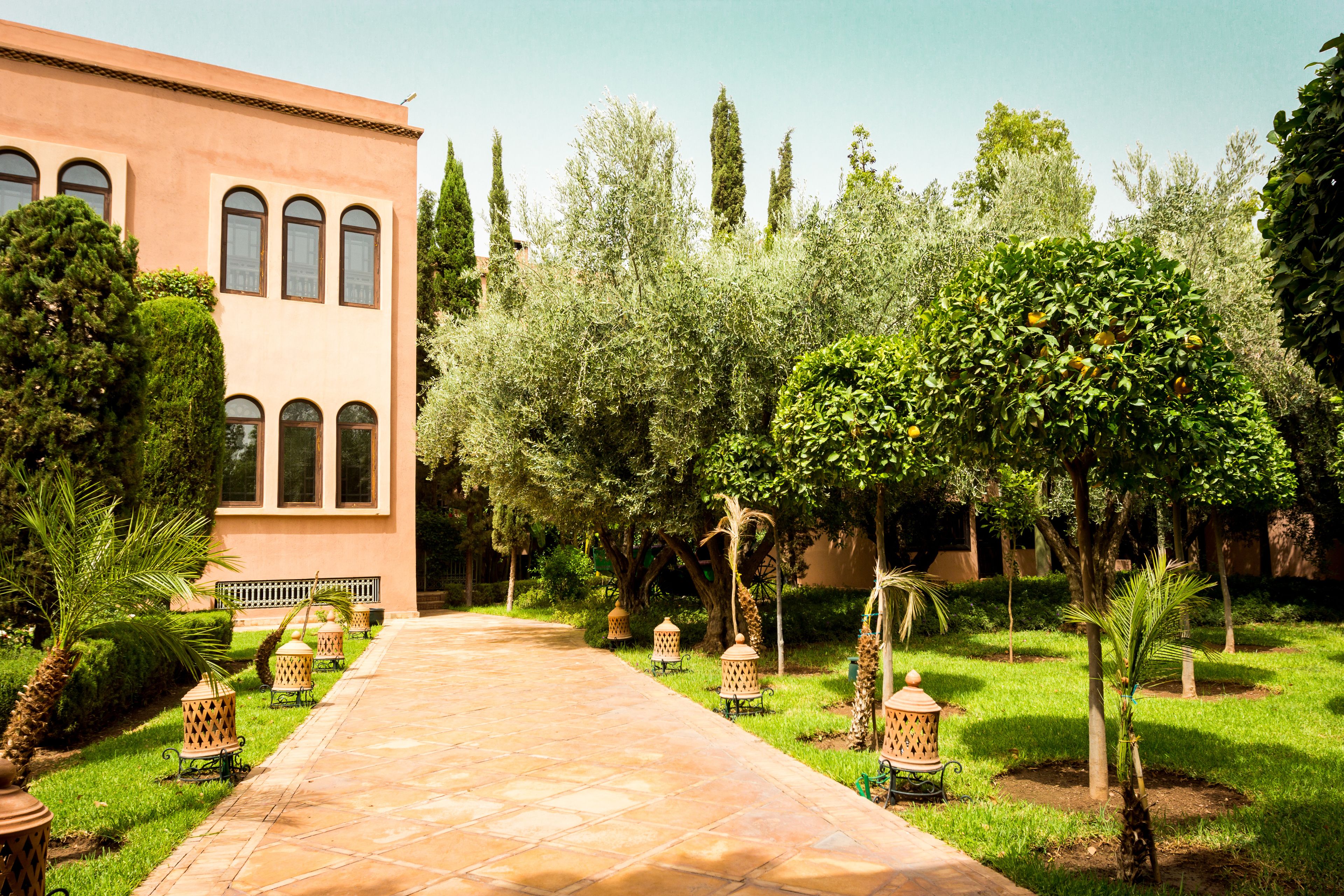 A paved pathway leading to a building with arched windows, surrounded by lush greenery and palm trees.