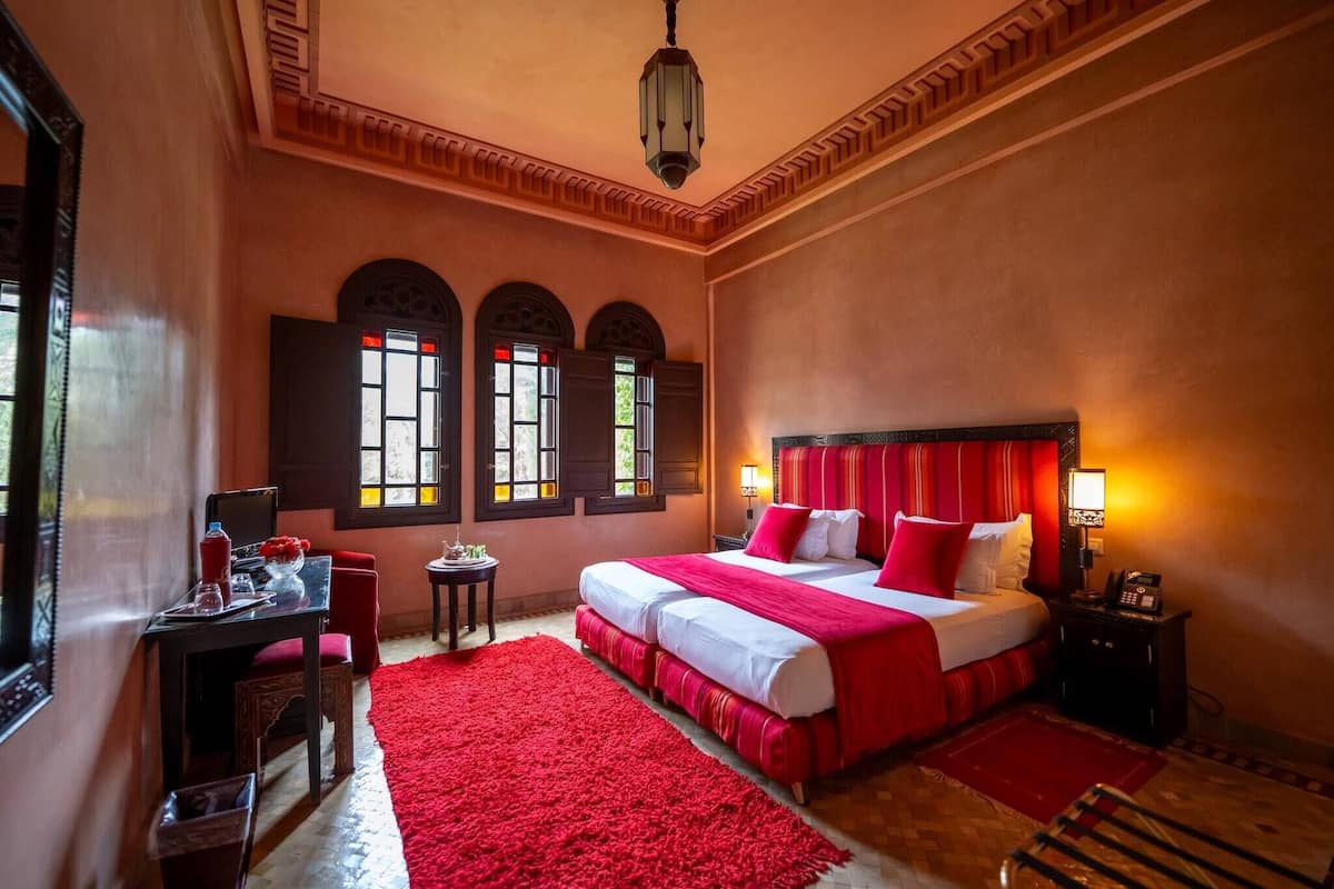 Quarto de hotel com uma cama grande, uma escrivaninha com cadeira, uma mesa pequena e um tapete vermelho.