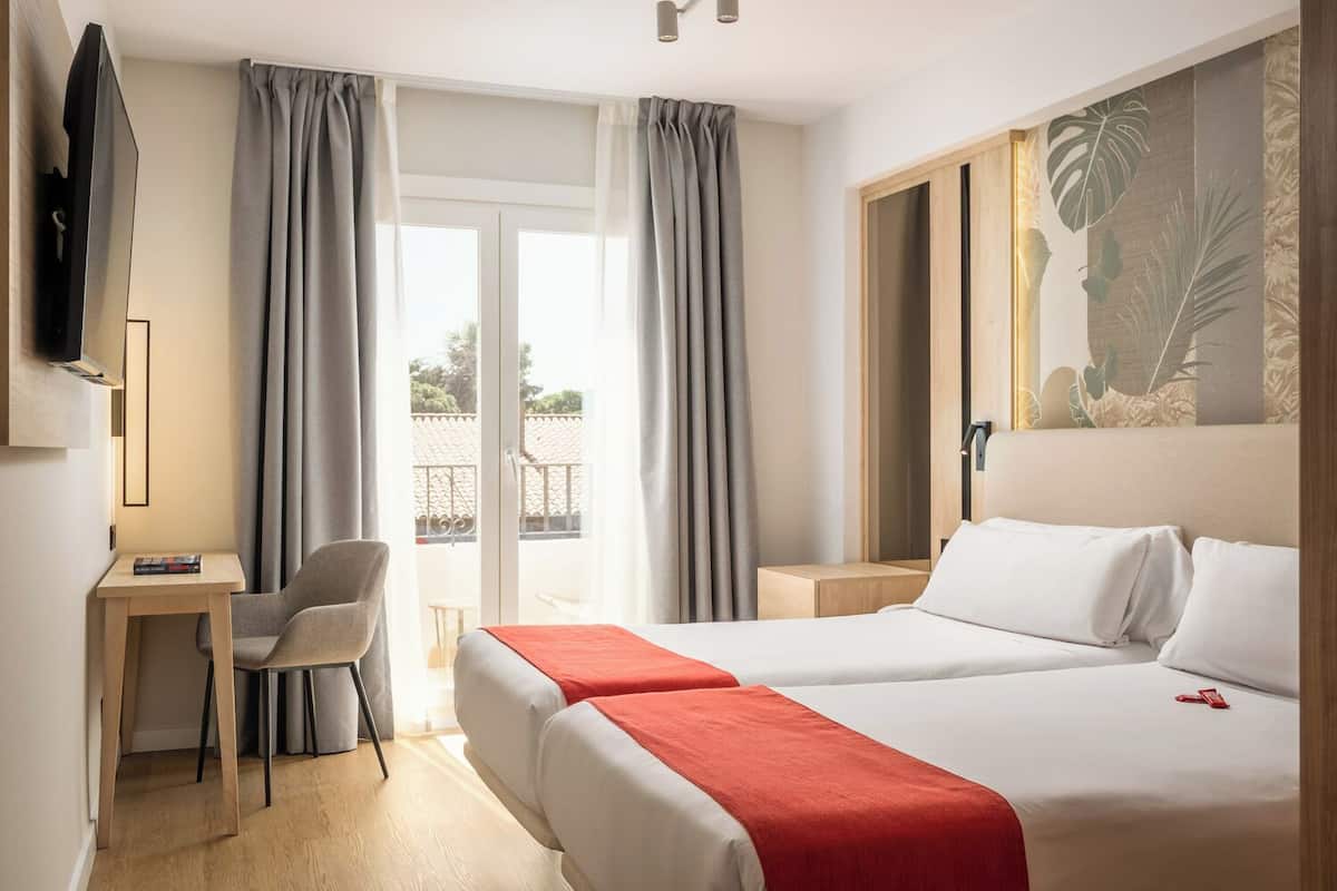 Une chambre d’hôtel avec deux lits, un bureau, une chaise, une télévision et un balcon avec des rideaux.