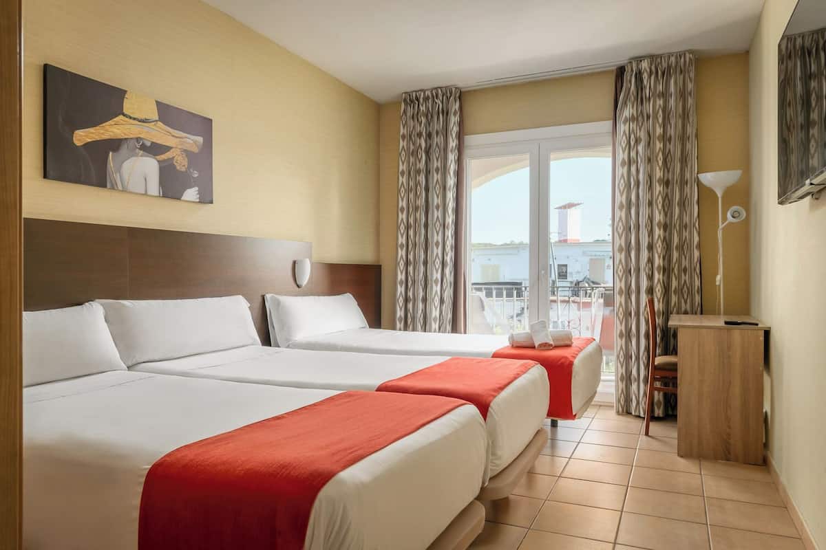 Une chambre d’hôtel avec deux lits, un bureau, une chaise, un balcon avec des rideaux et un tableau au mur.