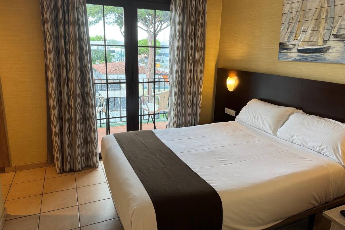 Une chambre d’hôtel avec un lit, des rideaux, une lampe fixée au mur et une vue sur l’extérieur.