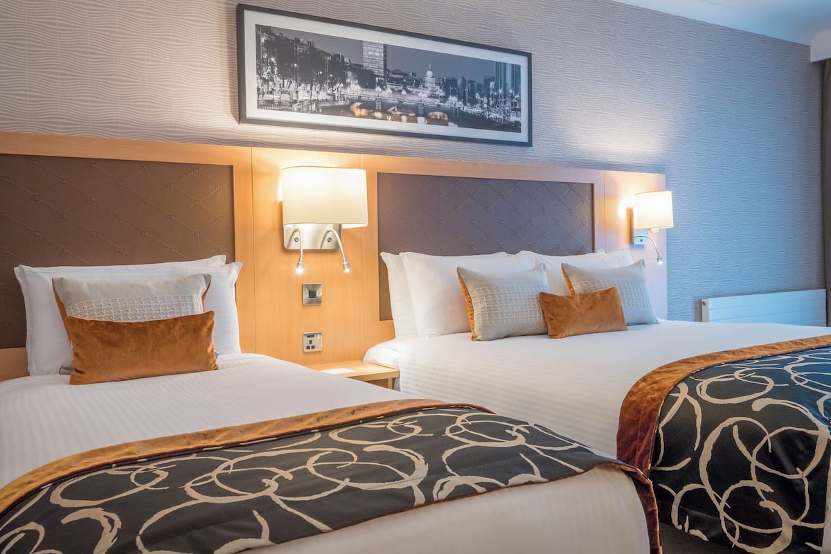 A hotel room with two beds, a cityscape photo on the wall, and wall-mounted lamps.