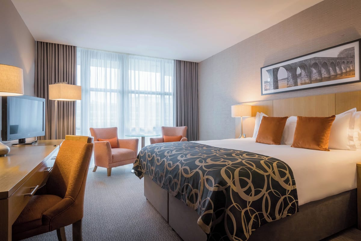 A hotel room with a large bed, a desk with a TV, two chairs, and a window with curtains.