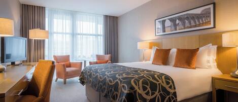 A hotel room with a large bed, a desk with a TV, two chairs, and a window with curtains.