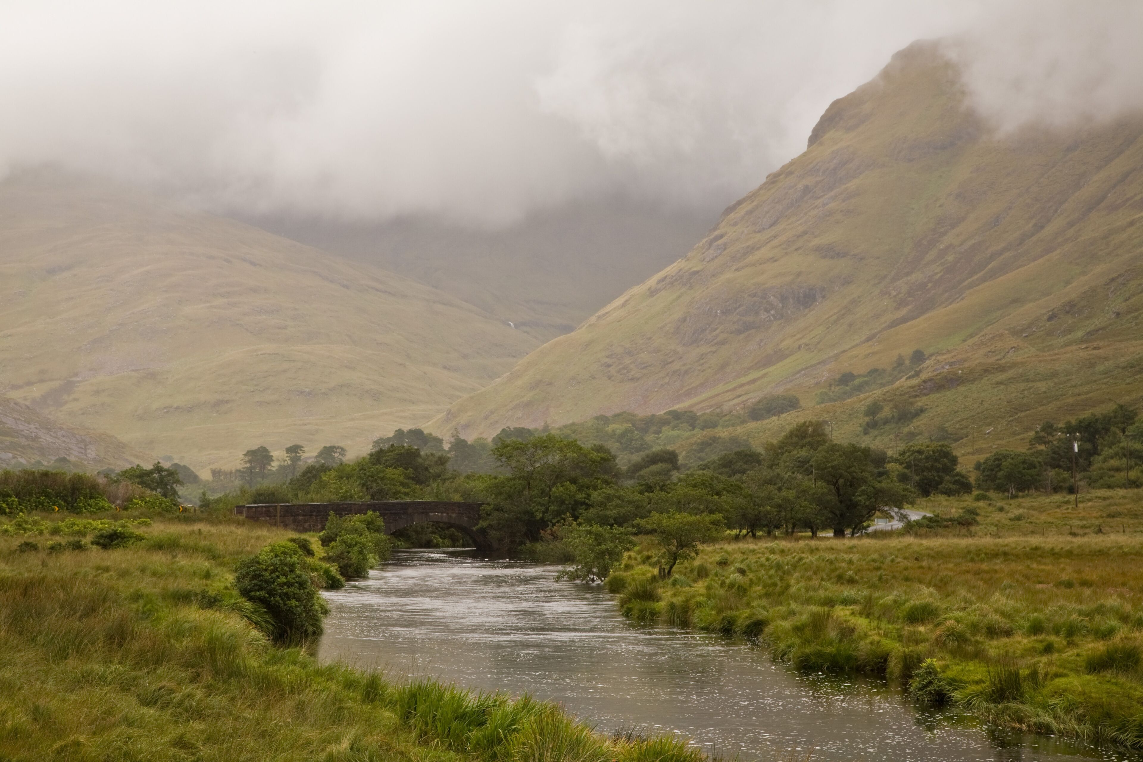 Une rivière qui traverse une vallée, avec un pont et des collines en arrière-plan.