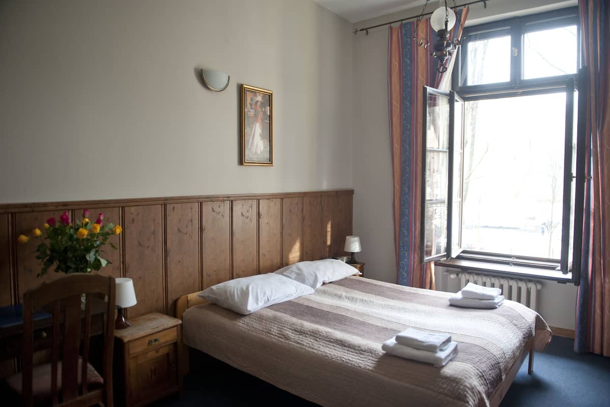 Una cama bien hecha con almohadas y toallas, un cabecero de madera, una mesita de noche con lámpara, y una ventana con cortinas.