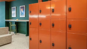 Une rangée de casiers orange, numérotés de 01 à 12, une chaise verte, une petite table sur laquelle se trouve un objet jaune, et deux tableaux encadrés sur des murs de couleur turquoise.