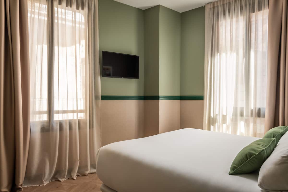 A hotel room with a bed, green walls, a mounted TV, and sheer curtains.