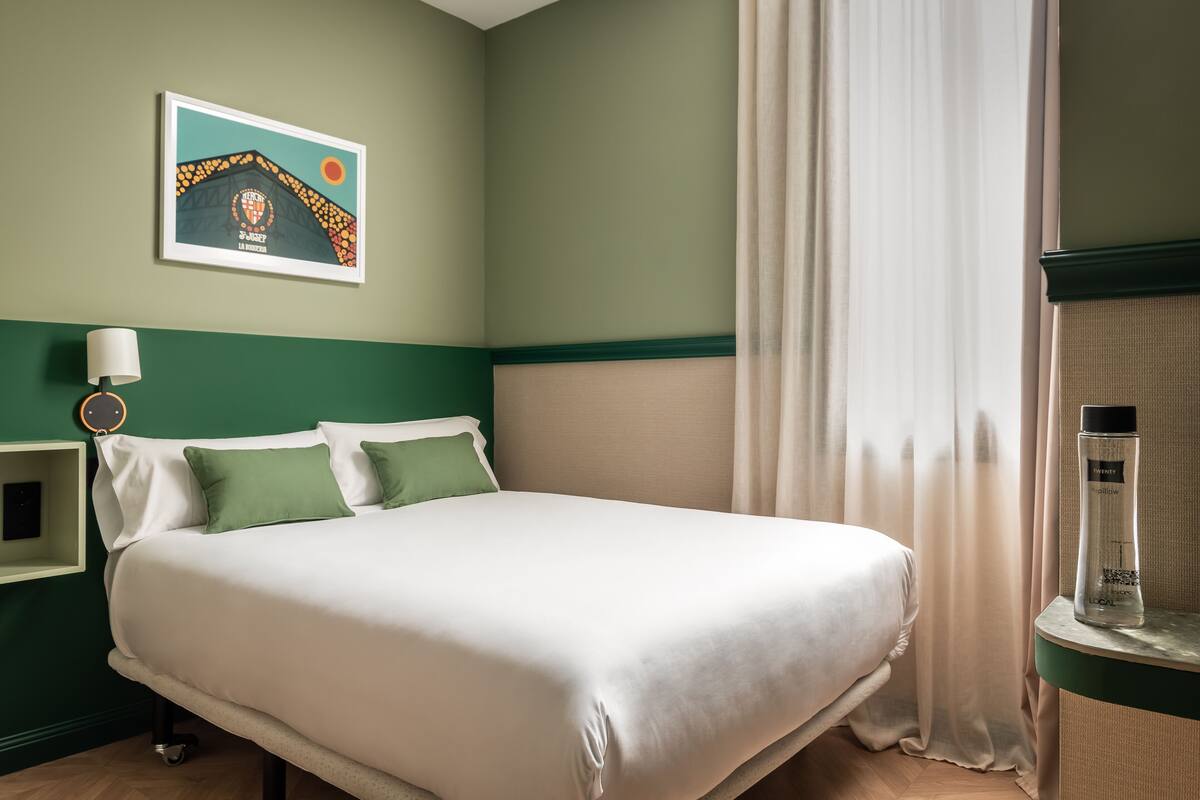 A neatly made bed with white bedding and green pillows, a framed picture on the wall, a bedside table with a lamp, and a bottle of lotion on a nightstand.