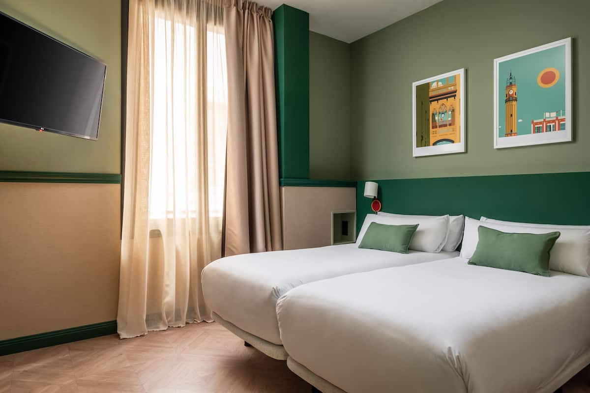 A hotel room with two beds, a flat-screen TV, a window with curtains, and two framed pictures on the wall.