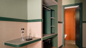 Une petite salle de bain aux couleurs vertes et beiges, avec un plan de toilette sur lequel on aperçoit une bouteille d’eau et des verres, un petit lavabo et des toilettes visibles à travers une porte ouverte.