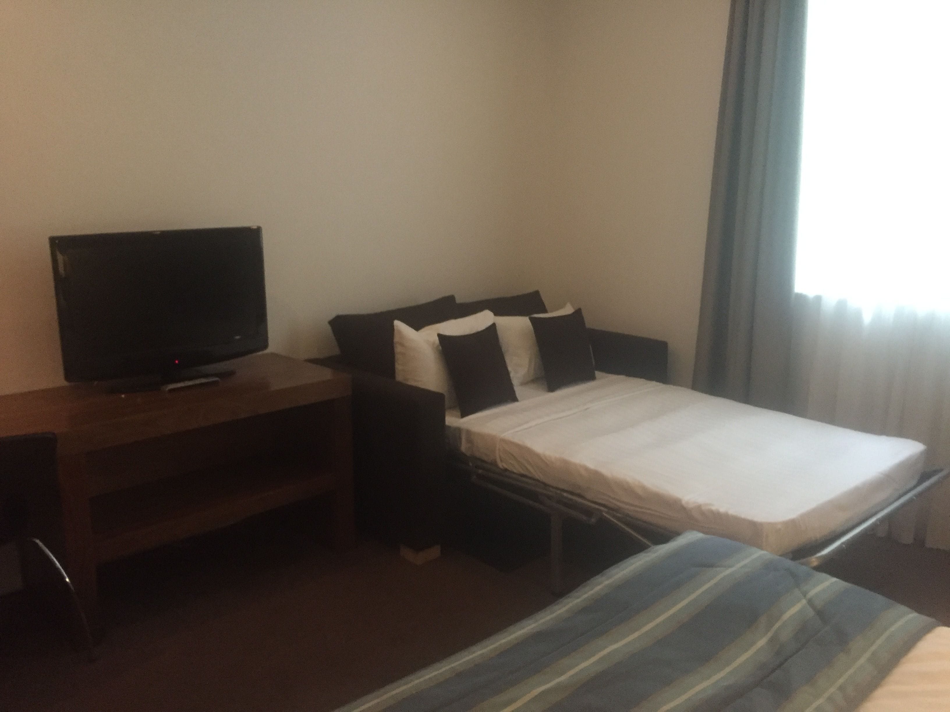 Una habitación de hotel pequeña y sencilla, con una cama, un televisor sobre un soporte de madera y una ventana con cortinas.