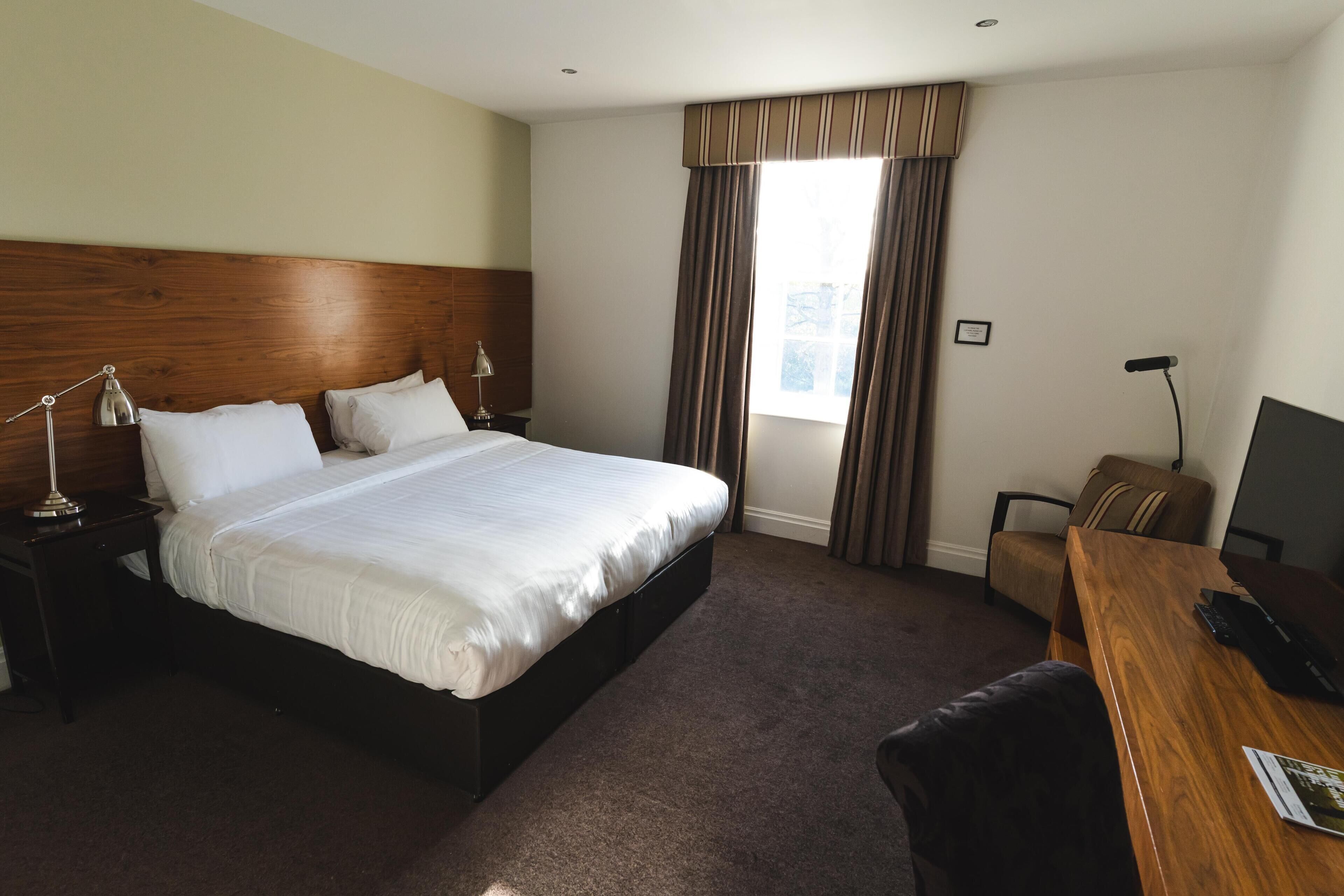 A hotel room with a large bed, a desk, a chair, a lamp, and a window with curtains.