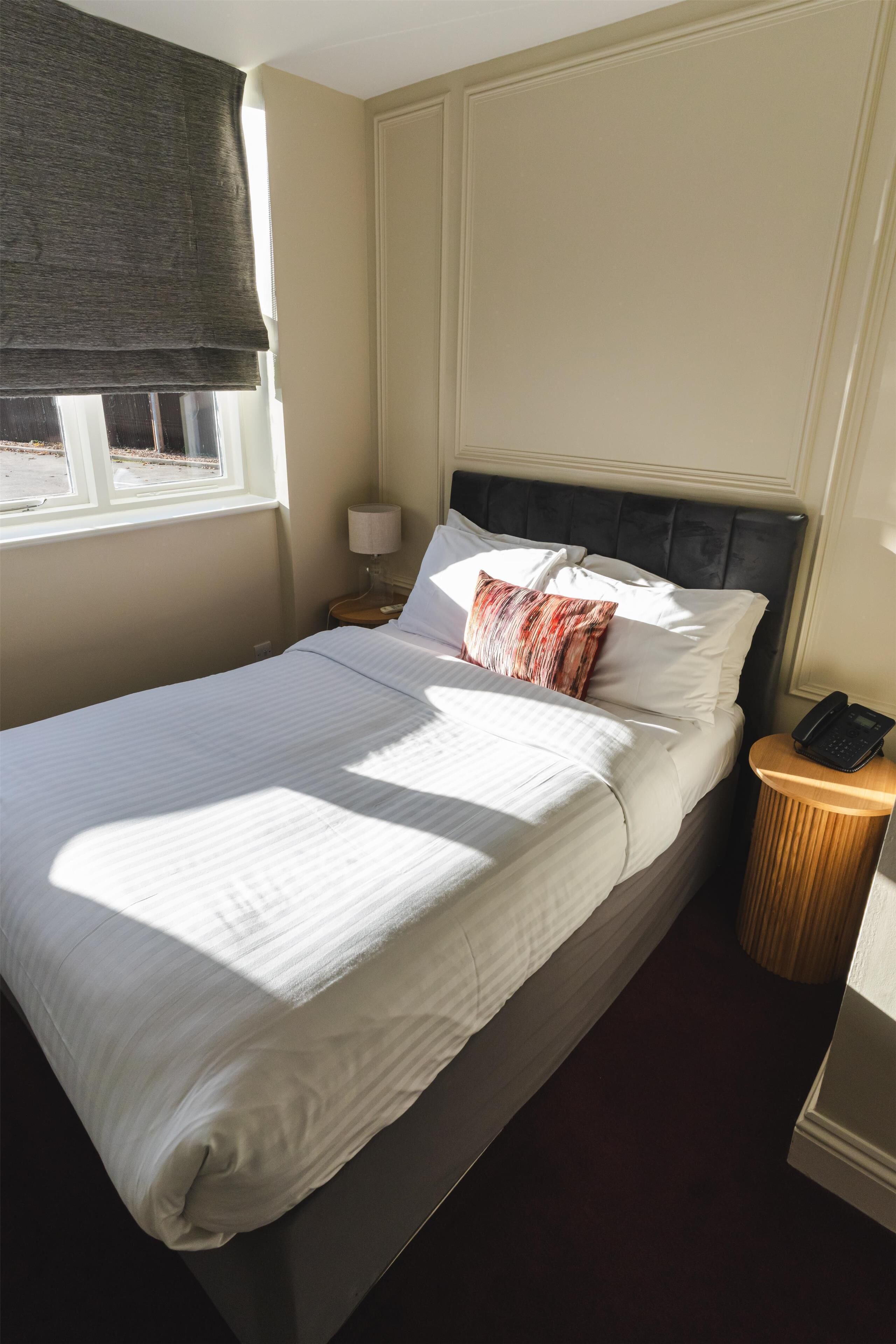 A neatly made bed with white linens, a dark headboard, a bedside lamp, and a telephone on a wooden nightstand.