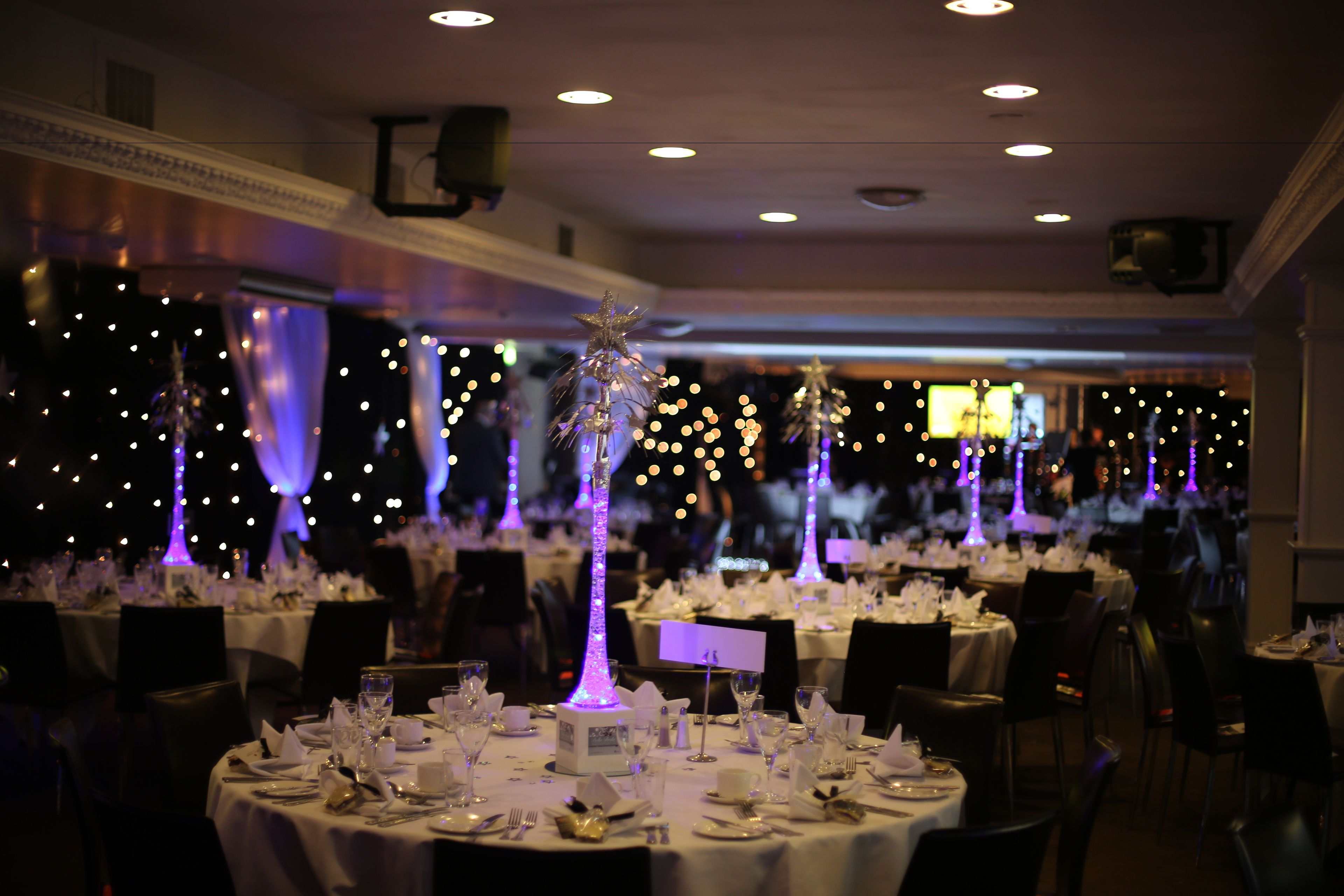 Un salón de banquetes con mesas redondas dispuestas para un evento formal, con centros de mesa iluminados y una iluminación ambiental tenue.