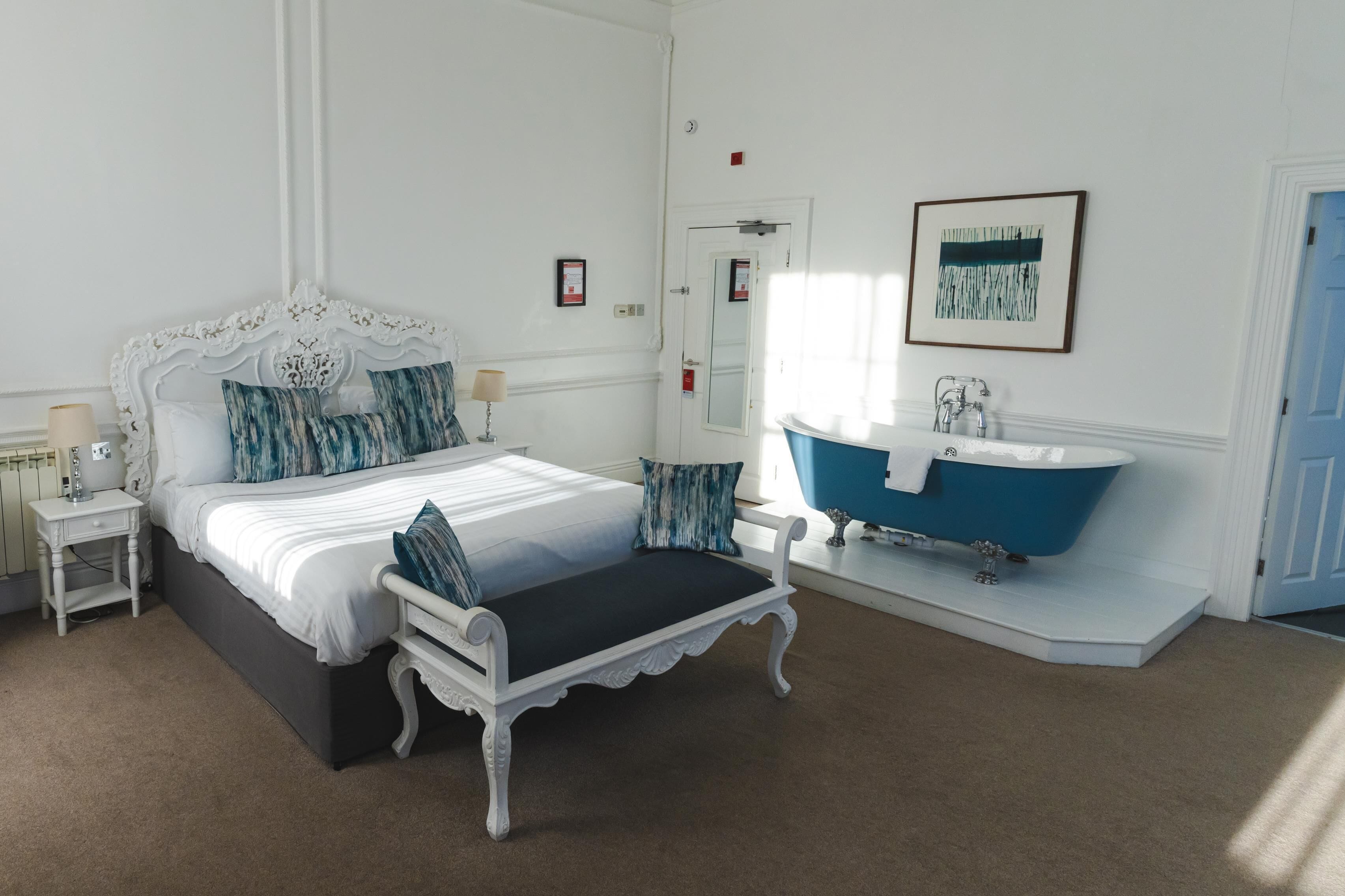 A bedroom with a large bed, a nightstand, a vanity with a blue bathtub, and a door with a mirror.