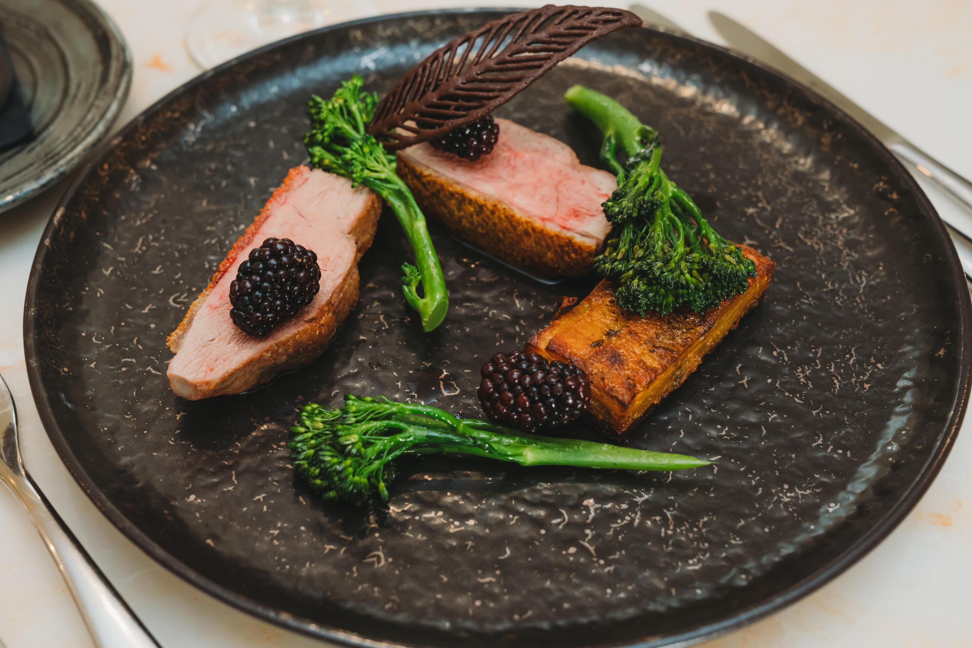 A plate with slices of meat, broccoli, and blackberries.