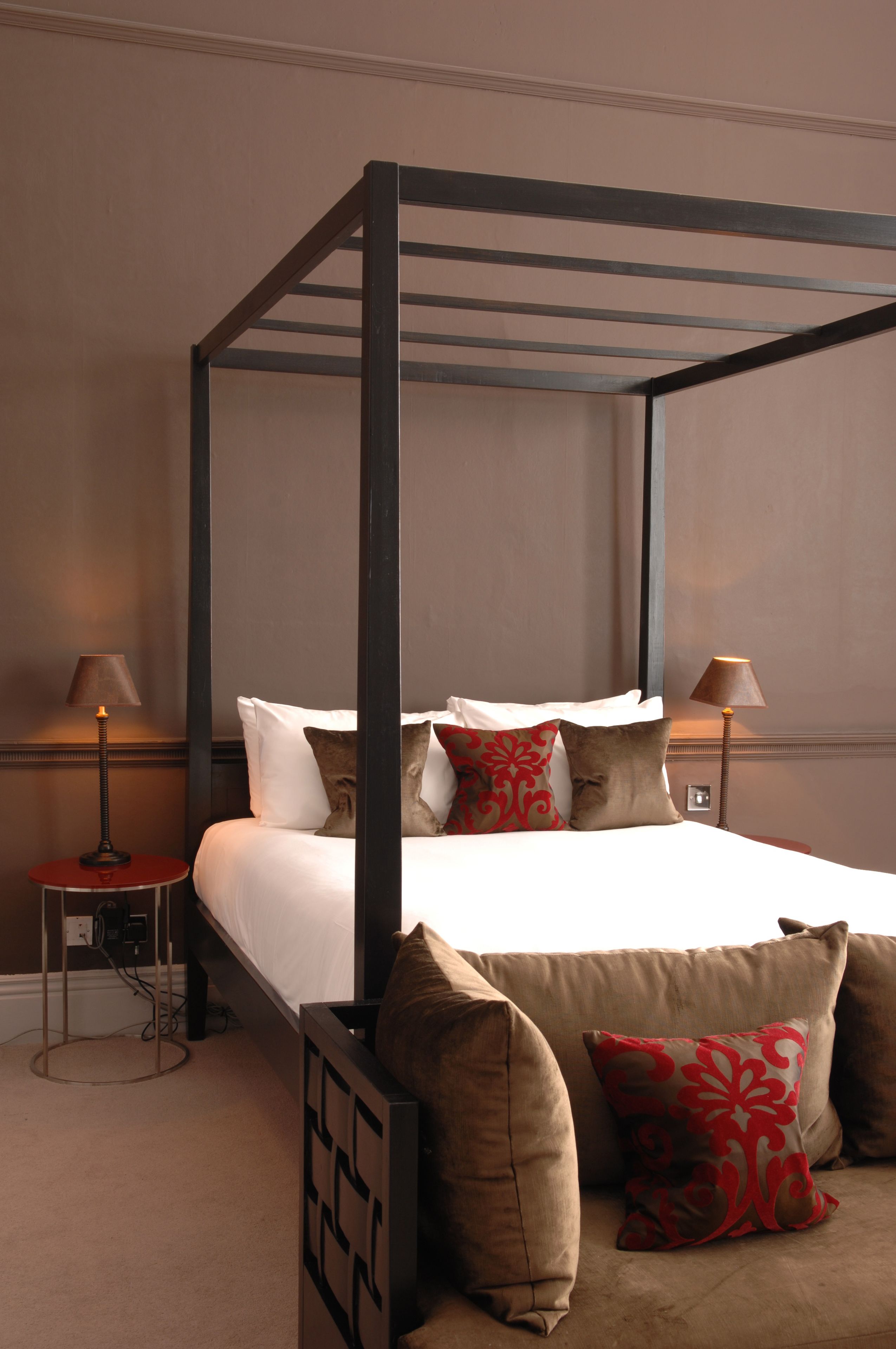 A four-poster bed with white linens and multiple pillows, a bedside table with a lamp, and a wall-mounted electrical outlet.