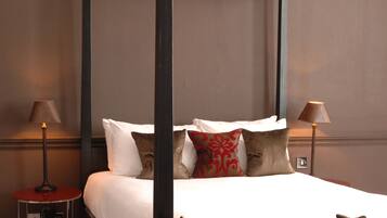 A four-poster bed with white linens and multiple pillows, a bedside table with a lamp, and a wall-mounted electrical outlet.