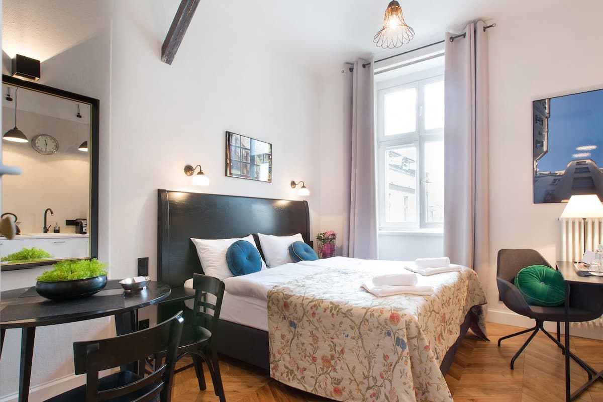 Una habitación de hotel moderna con una cama grande, una mesa de comedor y sillas. Cuenta con una ventana con cortinas, un reloj en la pared y un espejo.