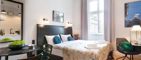 Una habitación de hotel moderna con una cama grande, una mesa de comedor y sillas. Cuenta con una ventana con cortinas, un reloj en la pared y un espejo.