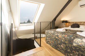 Een slaapkamer met dakraam, een bed met bloemenbeddelsels, een nachtkastje met lamp, en uitzicht op een gebouw door het raam.