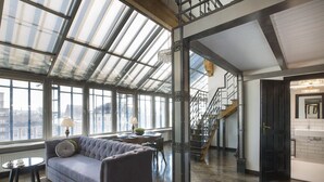 Ein geräumiger Wohnbereich mit hohen Decken, großen Fenstern, einem grauen Sofa und einer Treppe zu einer oberen Etage.