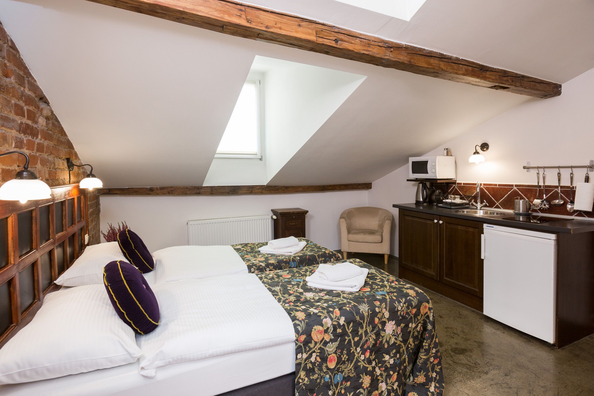 A cozy attic bedroom with a sloped ceiling, a skylight, a bed with white linens, a small kitchenette, and a wooden headboard.