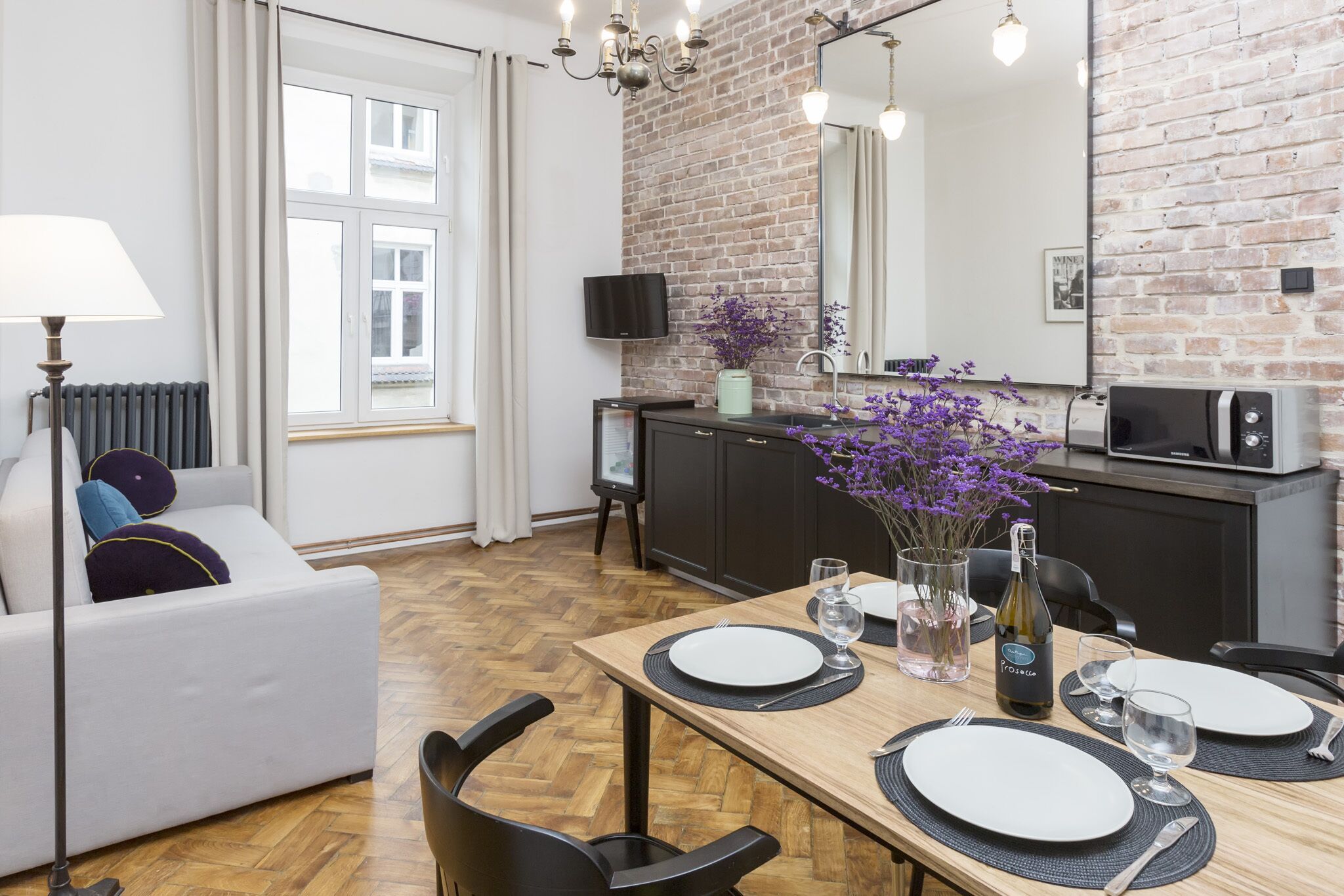 A modern living room with a sofa, a dining table set for two, a kitchen area with a microwave, and a brick wall.