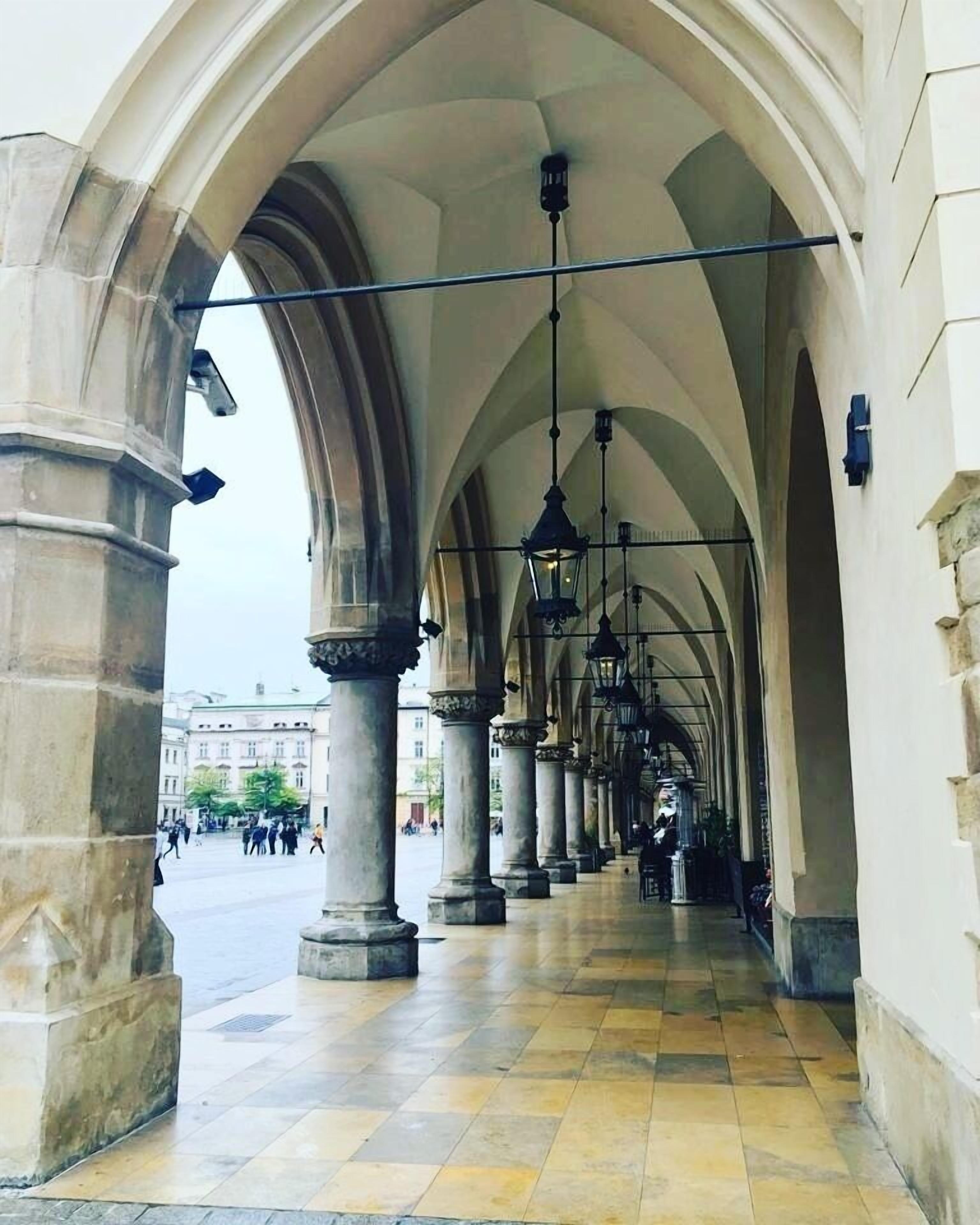 Ein Korridor mit Bögen und hängenden Laternen, der zu einem offenen Platz mit Gebäuden im Hintergrund führt.