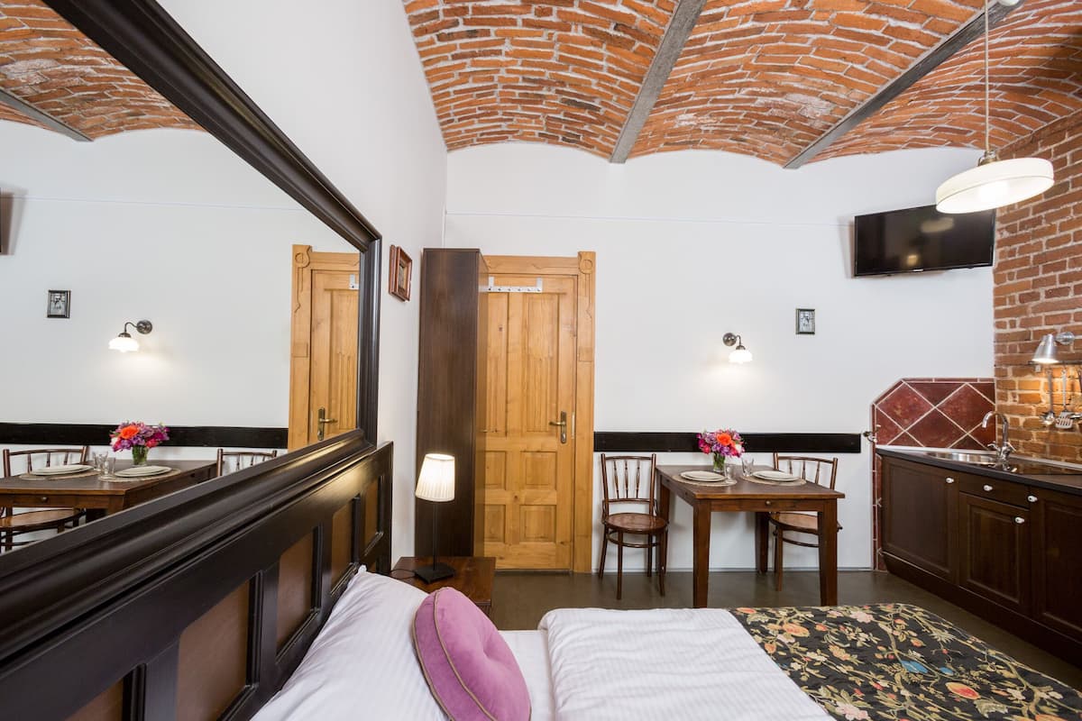 Una habitación de hotel con techo de ladrillo, una puerta de madera, una mesa de comedor con sillas, una cama con colcha estampada y un televisor montado en la pared.