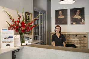 Een receptiebalie met een vaas rode bloemen, ingelijste portretten aan de muur, en een presentatie van boeken en een klok.