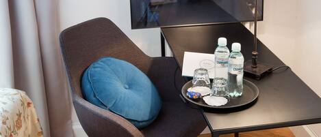 A hotel room with a single chair, a small table with a lamp, a tray with water bottles and snacks, and a view of buildings through a window.