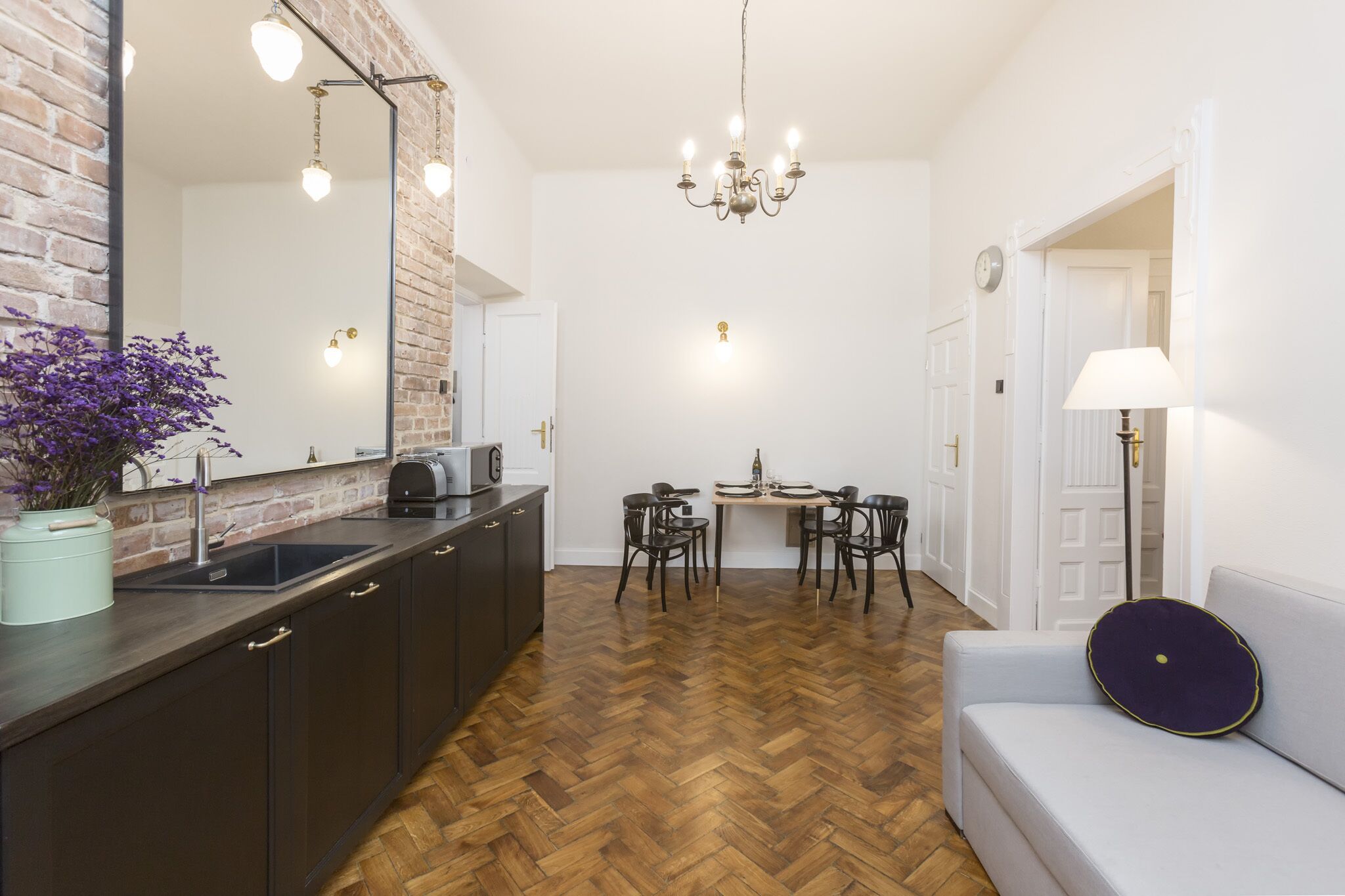 A compact kitchen with a herringbone floor, a dining area with four chairs, and a sofa with a decorative pillow.