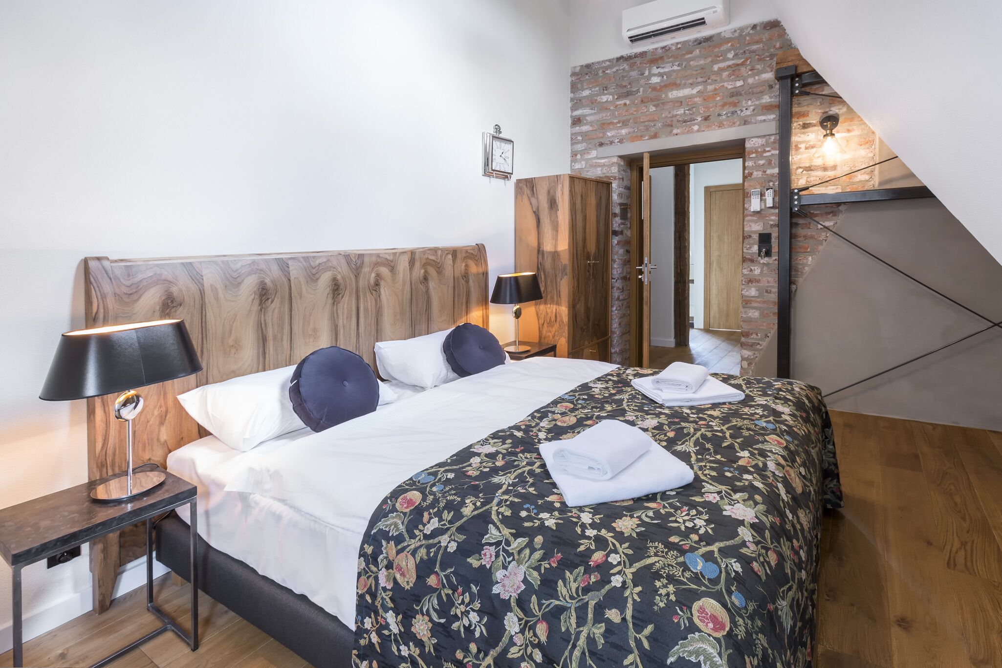 A bedroom with a wooden headboard, a bed with a floral bedspread, two bedside lamps, and a brick wall.