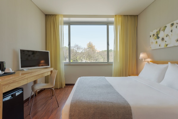 A hotel room with a bed, a desk with a television, a chair, and a window with curtains. - HF Fénix Garden (Lisbon)