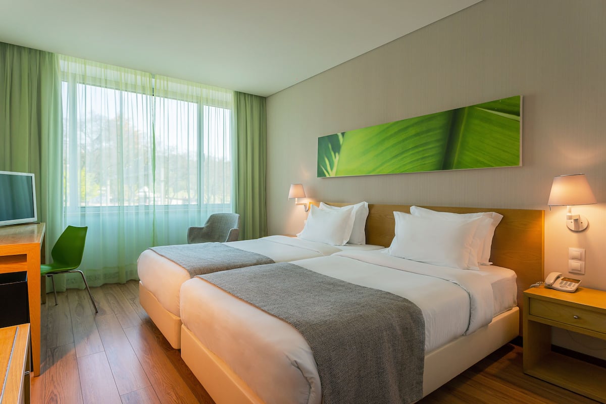 Habitación de hotel con dos camas, un escritorio, una silla y un ventanal con cortinas translúcidas.
