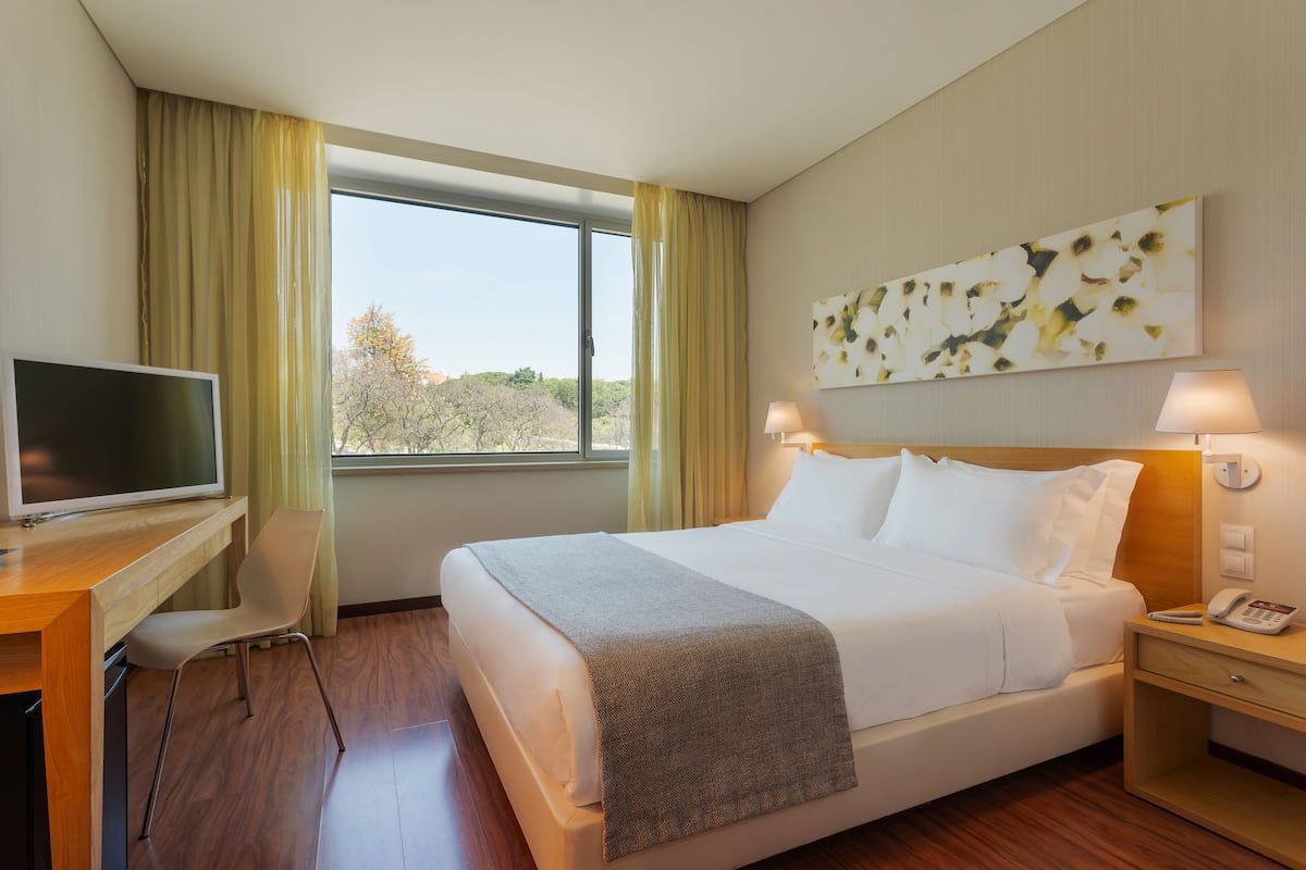 Una habitación de hotel con cama, escritorio con televisor, silla, mesita de noche con lámpara y ventana con cortinas.
